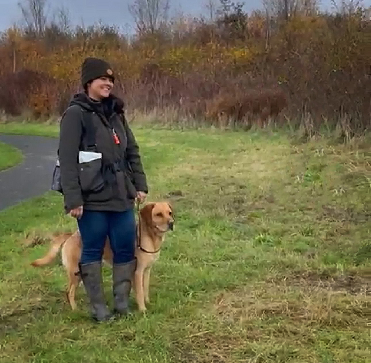 Eva staat glimlachend met haar labrador Lando aan de leiband in een herfstig buitengebied